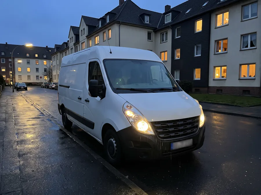 Ein weißer Transporter steht abends mit eingeschaltetem Licht in einer Wohnstraße.